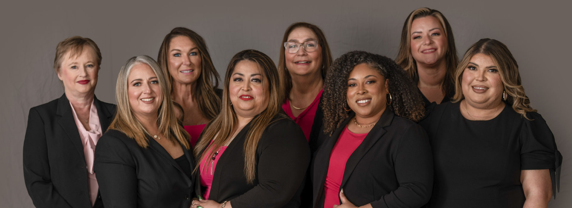 eight women in business attire standing and smiling in front of a gray background